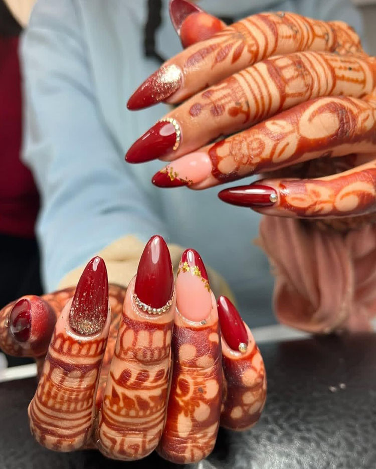 Detailed bridal nail art with deep red polish, gold leaf accents, and crystal embellishments paired with traditional henna hand designs